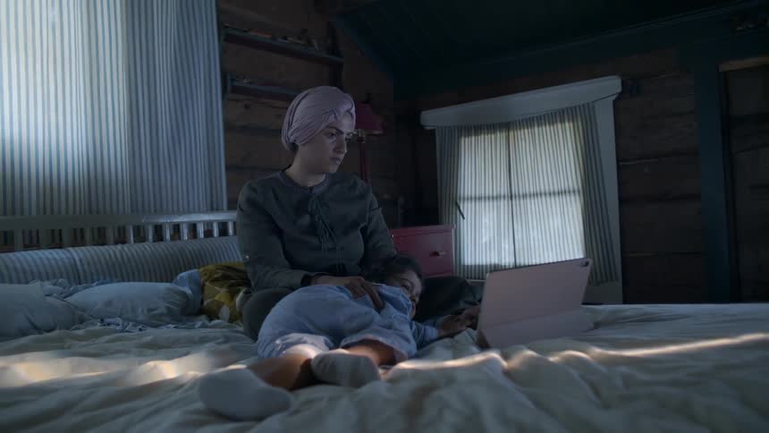 A mother and young daughter use a tablet in bed
