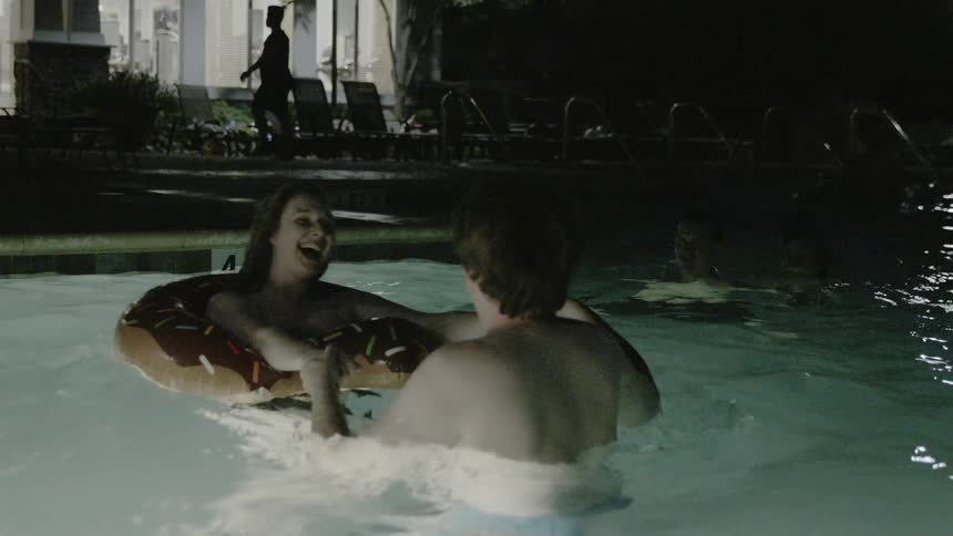 A man and woman smile while spinning in an outdoor swimming pool at night