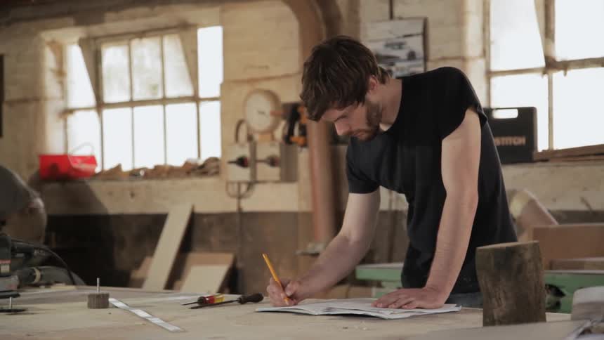 A man standing at a workbench makes notes and diagrams, another man joins him
