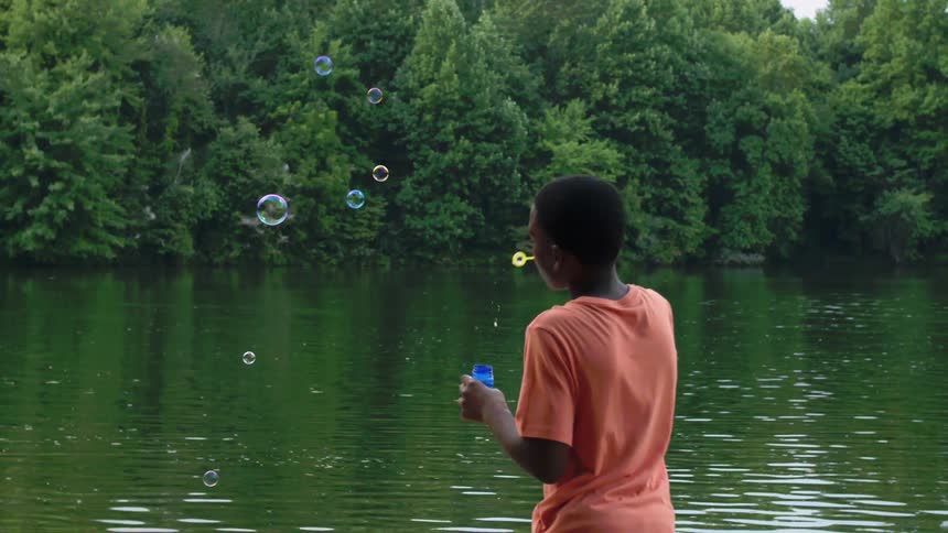 A boy stands beside a lake blowing bubbles