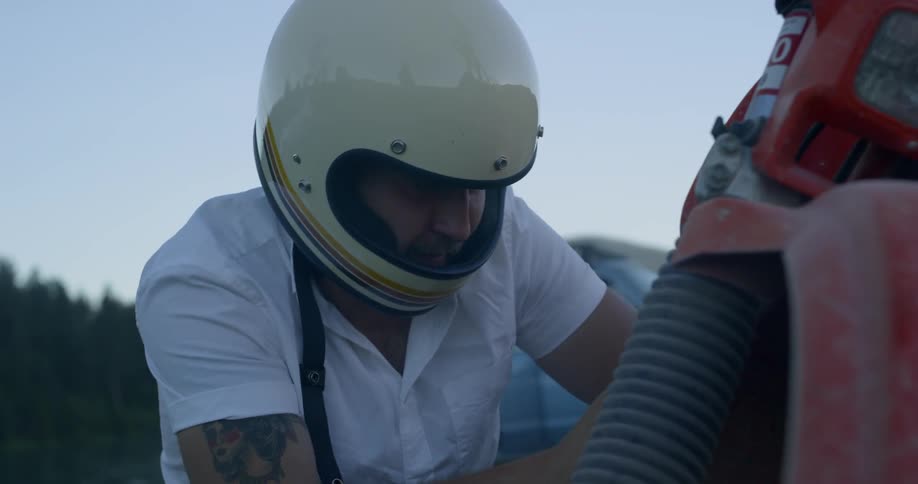 A man sitting next to an old motorbike in a white helmet fixing something on the bike