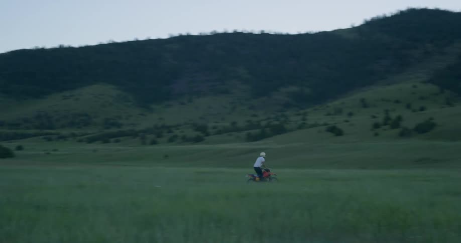 A person in a white helmet rides a red dirt bike through a field with mountains and evergreens in the background