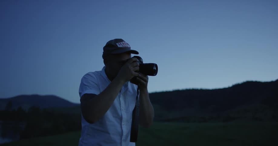 Man wearing cap and white shirt takes picture at dusk