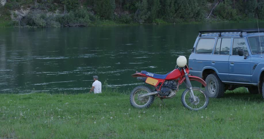 A man fishing in a river with an suv and motorbike nearby