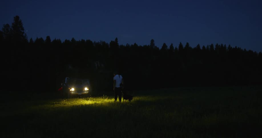 A man an a dog walking towards a car in a dark field at night