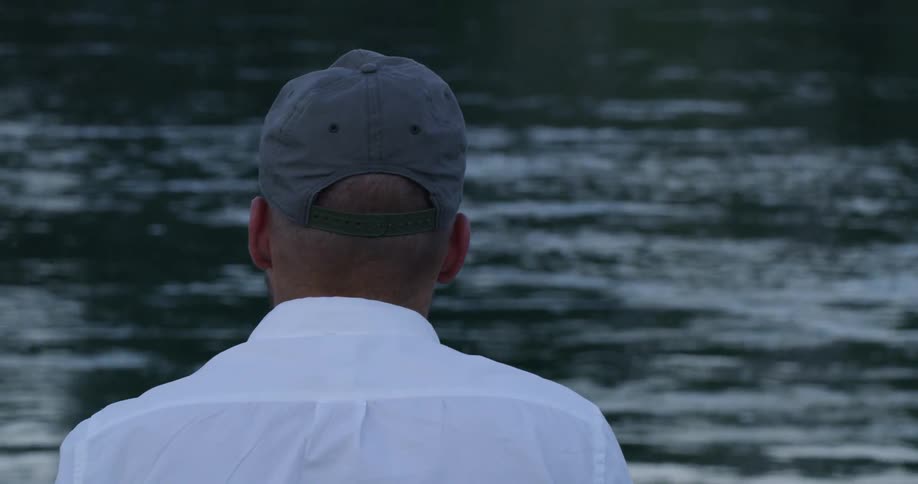 Man wearing cap and white shirt looks around by lake at dusk