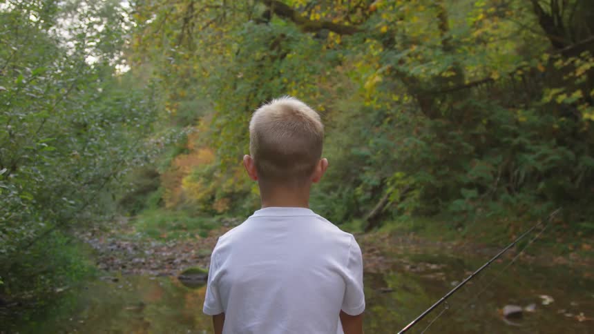 A young white boy is fishing and looking out at the forest beyond the water