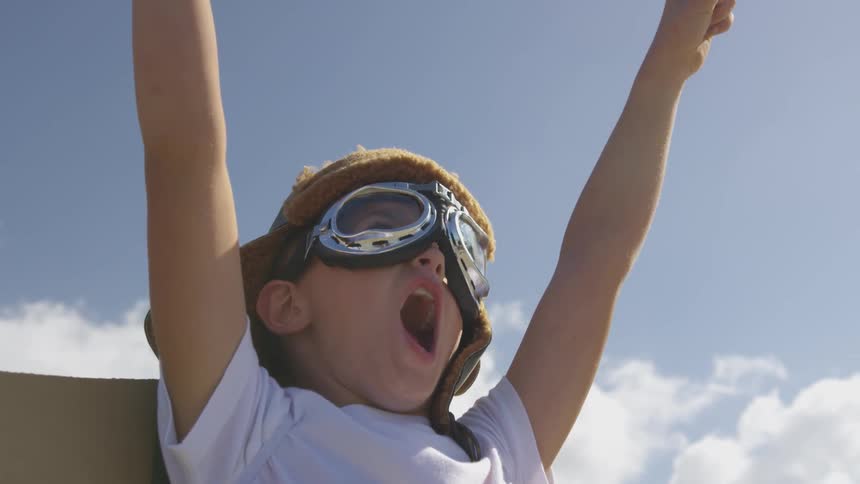 A young white boy wearing an aviator hat, goggles, and cardboard cape yells with his hands reached above him towards the sky