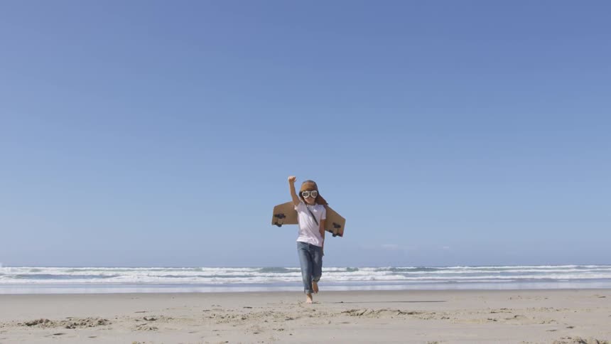 A little girls running on the beach with goggles and a airplane wings strapped to her