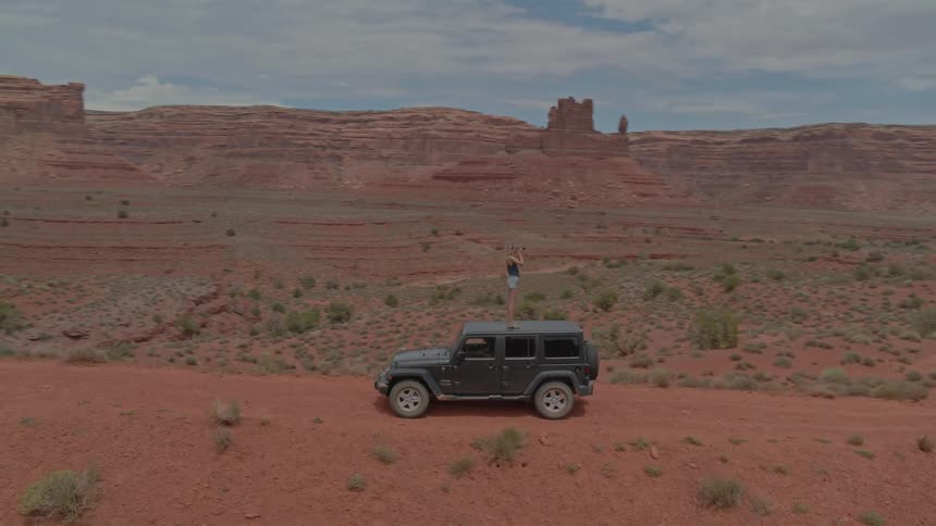 A woman in hot pants stands on a four wheel drive vehicle to take pictures from a dirt road in the desert