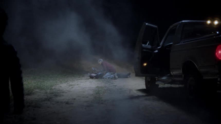 A man kneels next to a body on a dirt road at night