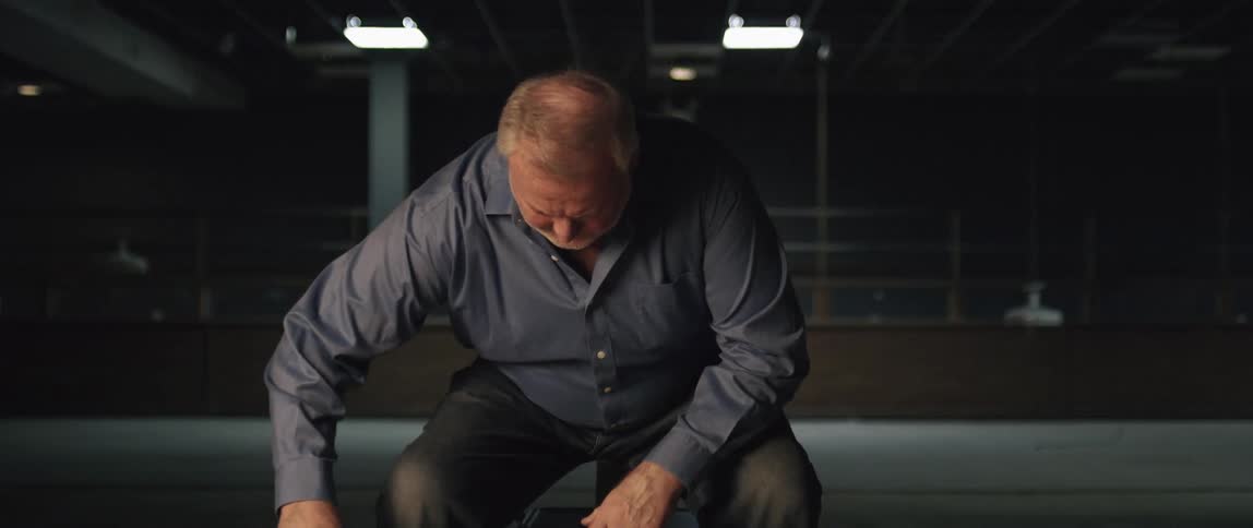 An elderly man walks towards a chair and sits down