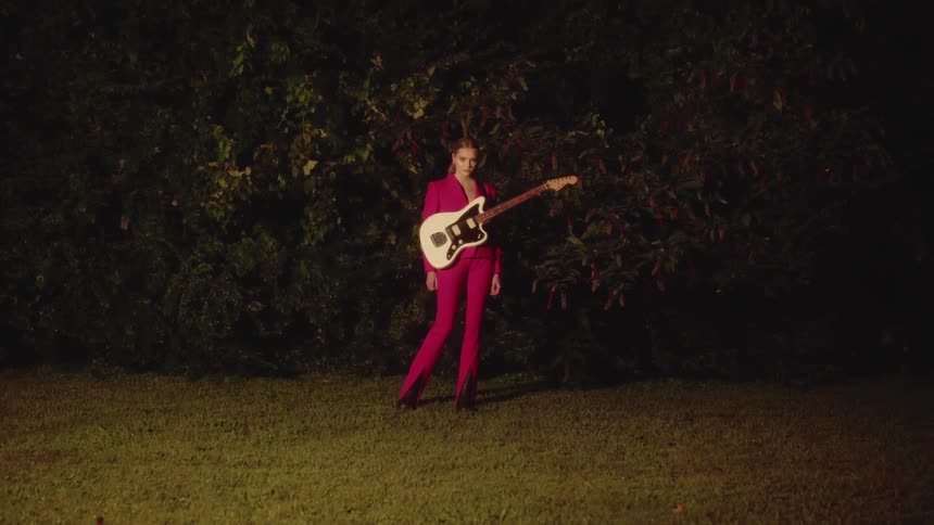 A woman in a pink suit stands outside with an electric guitar