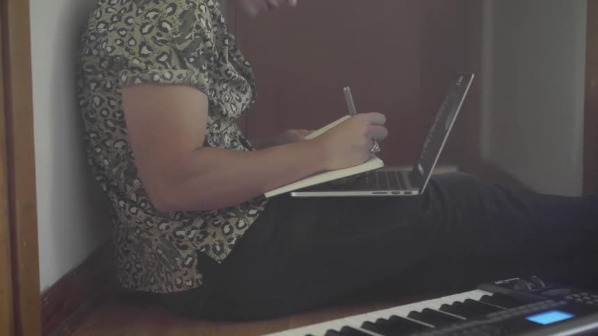 A man sits beside an electronic keyboard with a laptop on his lap as he takes notes