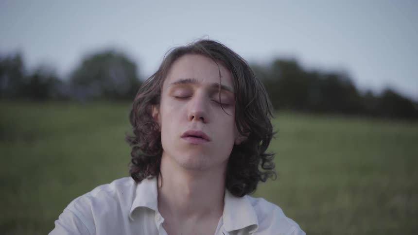 A young man sits in a grassy field with his eyes closed as if meditating