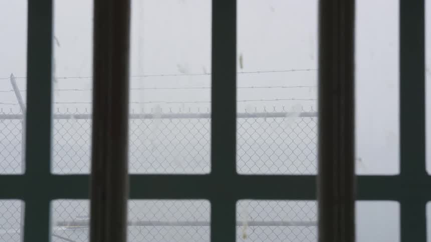 A tall prison fence with barbed wire is visible through a window with bars