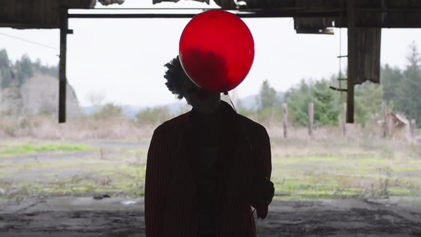 A clown stands in an empty building that is falling apart, holding a red balloon
