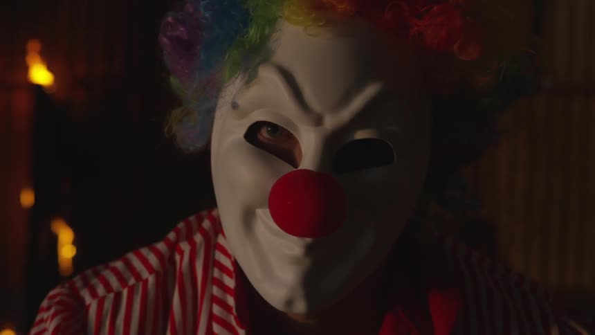 A fire burns behind a person in a clown mask