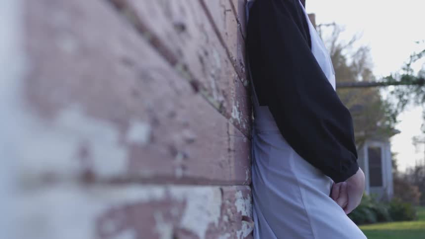 A lady wearing a white apron leans against a brick wall with peeling red paint