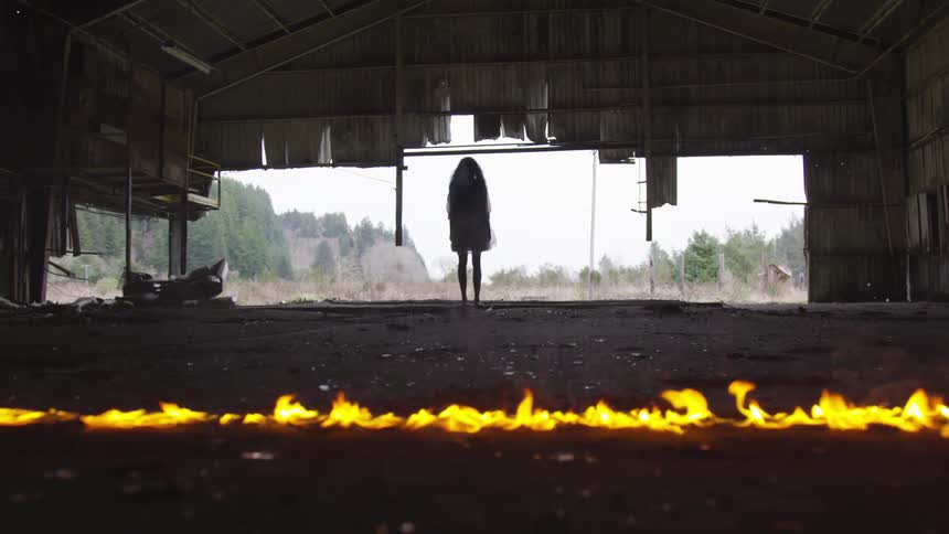 A woman stands in a shed while a fire burns behind her