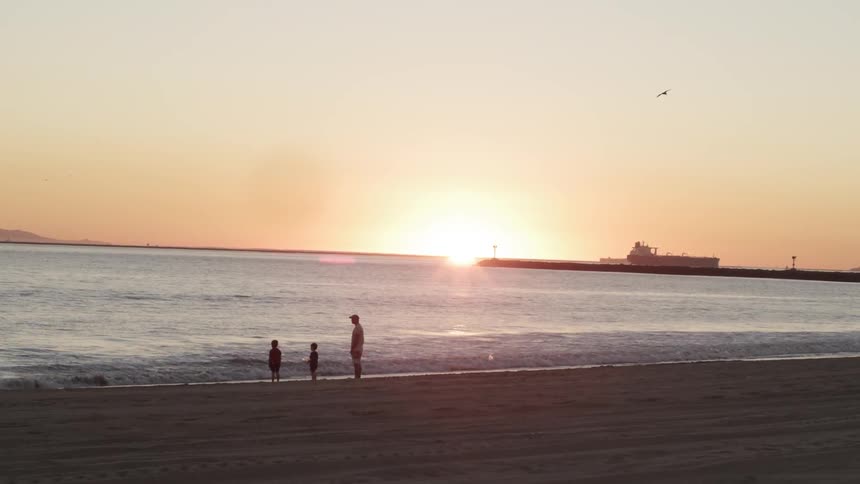 A man and two children stand alone on a beach watching the sun set