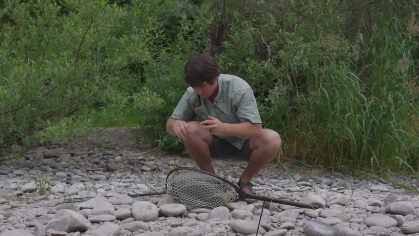 A man uses his teeth to break a fishing line while squatting on rocks in front of a forested area with a fishing net at his feet