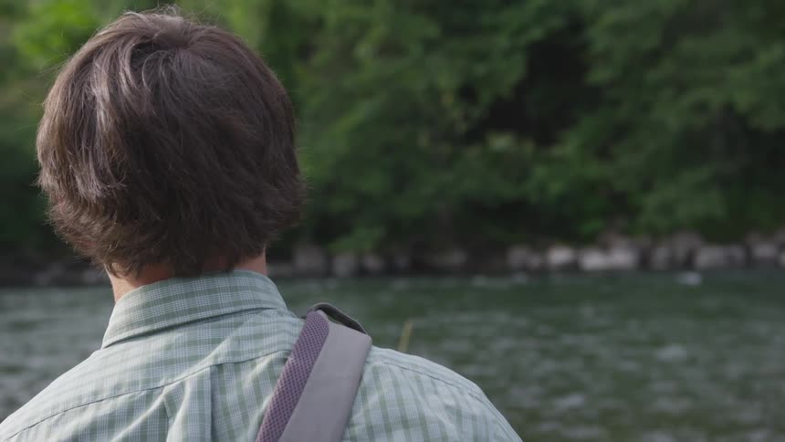 A man in a blue plaid buttoned shirt stands facing a dense forest on the other side of a river