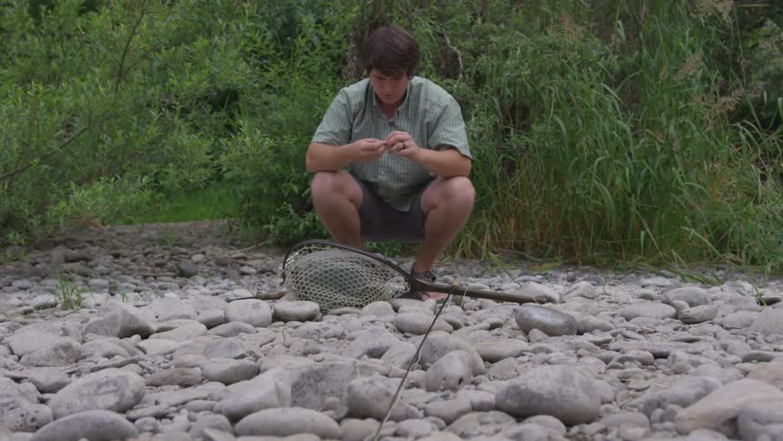 A man squats on the rocks of a riverbank baiting a hook