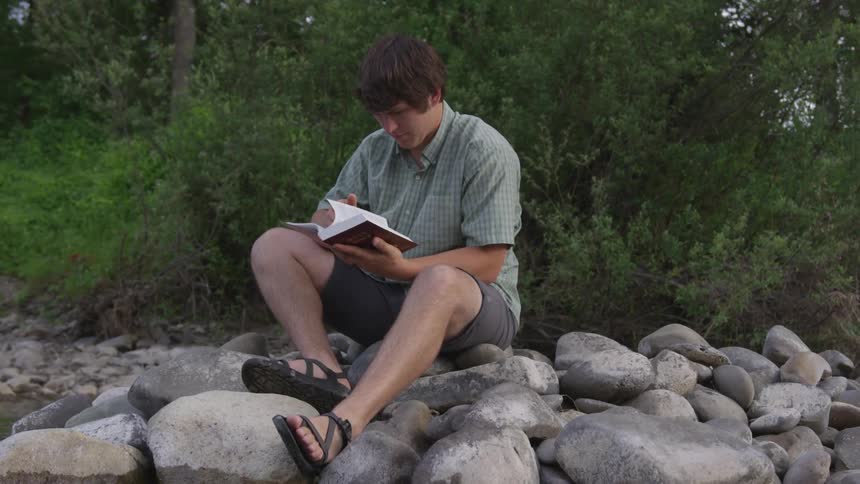 White man sitting at the rocks reading a book next to a lake