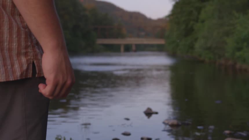 A man stands looking out over a river with forest on either side and a bridge in the distance