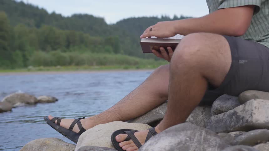 A thick legged man wearing sandals and shorts is sitting on some smooth rocks adjacent to a gently flowing river while turning pages in a book