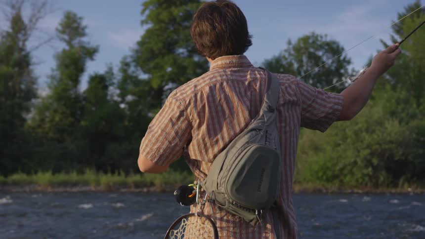 A man wearing a checkered shirt and backpack is standing in the stream throwing his fishing line in and out of the water