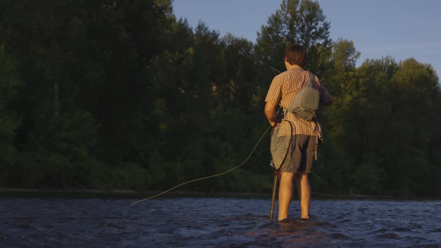 Man casts fishing line while standing in river across from woods