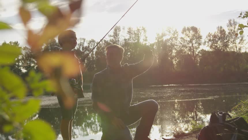 A father helps his son with a fishing pole next to a river at sunrise