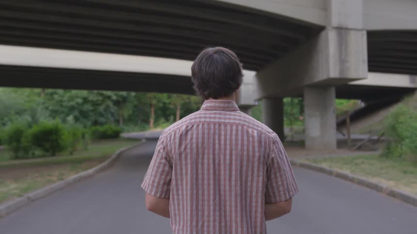 A young man walks on a concrete path under a bridge
