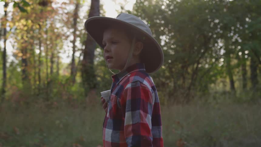 A small male child wearing a hat with a strap and holding a styrofoam cup is walking near the woods