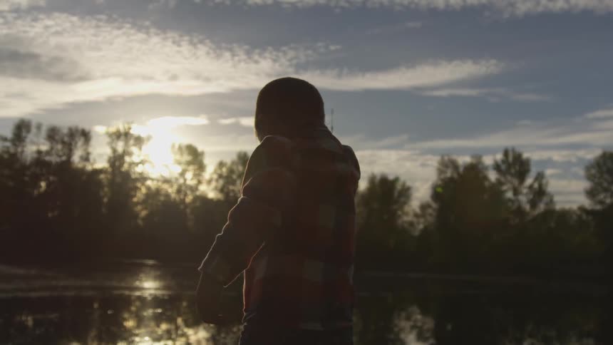 A young boy is standing at the edge of a river while he is checking his fishing line as he fishes in the river