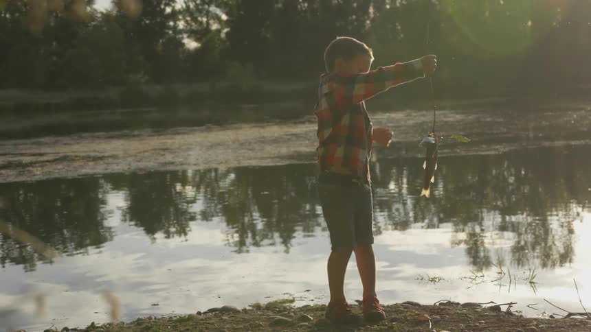 A young boy is standing on the dirt covered bank of a river as he holds up a fish that he just caught while it is still hanging on the fishing hook