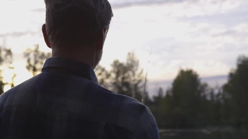 A man in a flannel shirt is motionless as he uses a fishing rod with a fishing line in a forested area