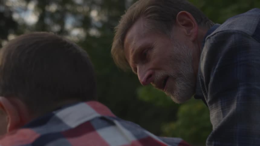 An older man with a beard is learning over and speaking words of advice to a young boy in a forested area