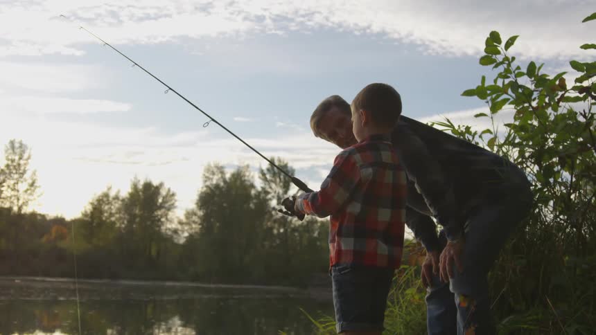 A dad leans down and smiles at his young son reeling in his fishing line