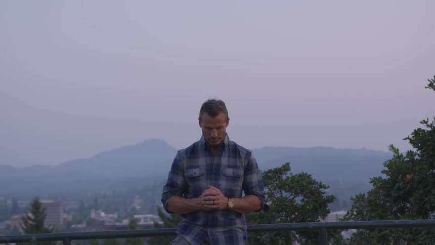 A white man in a blue flannel stands at a railing of an overlook with his head bowed in prayer