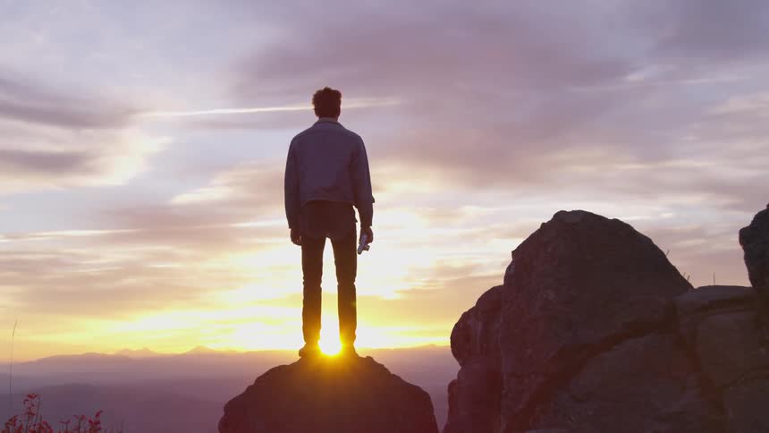 A man stands on a rocky ridge looking out at the sunset