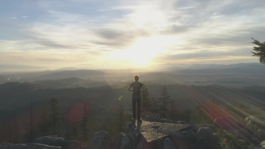 A man carrying a backpack stands on a large rock on top of a mountain looking out over the horizon