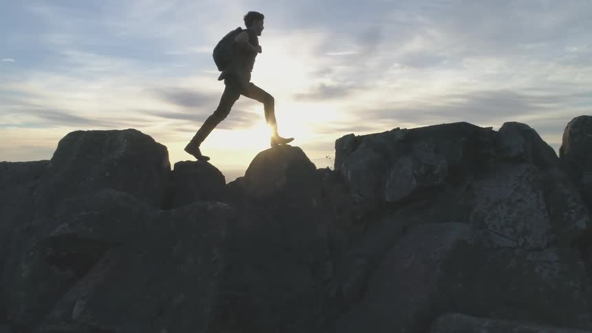 A tall man wearing a backpack hikes along a rocky ridge located above a gorgeous valley