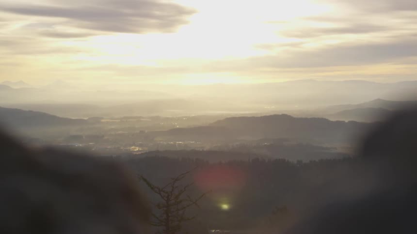 The sun is low in the cloudy sky as a valley can be seen from atop a mountain