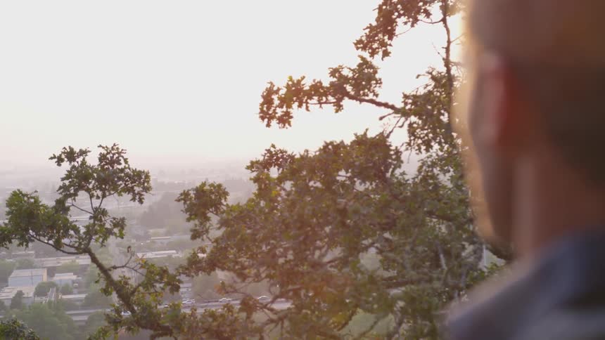 A man looks out from the top of a hill at sunset to an urban environment below