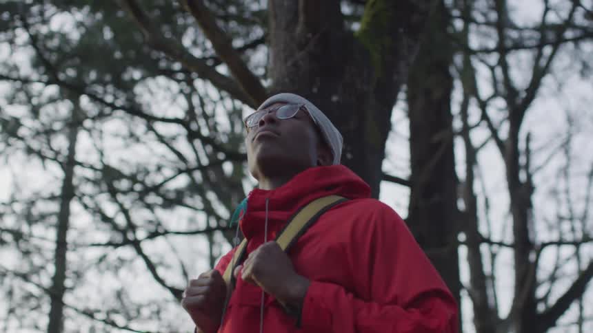 A black man carrying a backpack and wearing a skull cap eyeglasses and red jacket standing among trees while gazing around himself