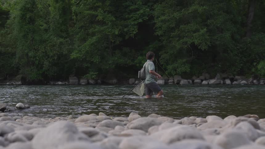 Man walks slowly through river as he carries fly fishing equipment