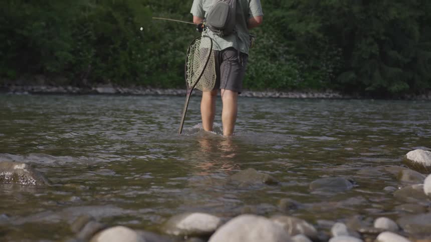 A man carrying fishing equipment walks on a shallow river